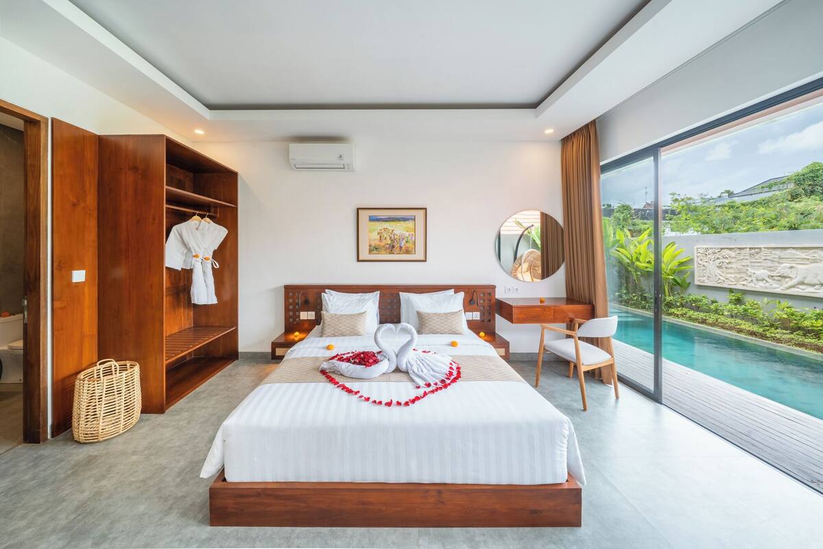 Ein modernes Hotelzimmer mit einem großen Bett, einem Holzschrank, einem Nachttisch, einem Wandgerät zur Klimatisierung und einem großen Fenster mit Blick auf Grünflächen.