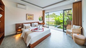 A modern hotel room with a large bed, a sitting area with a wicker chair, and a view of lush greenery through a sliding glass door.
