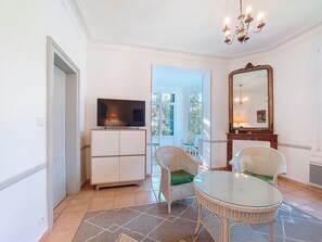 Une pièce comprenant un téléviseur posé sur un meuble, des fauteuils en osier, une table en verre, un lustre et un grand miroir.