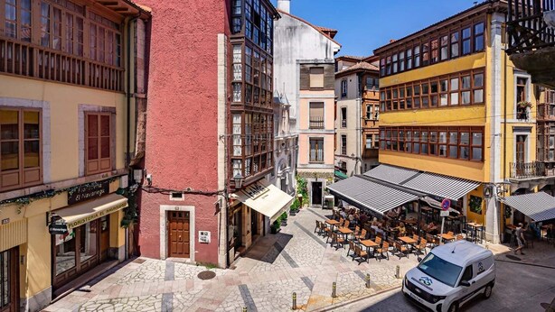 Una calle con edificios, un café con terraza y una furgoneta blanca estacionada al costado.