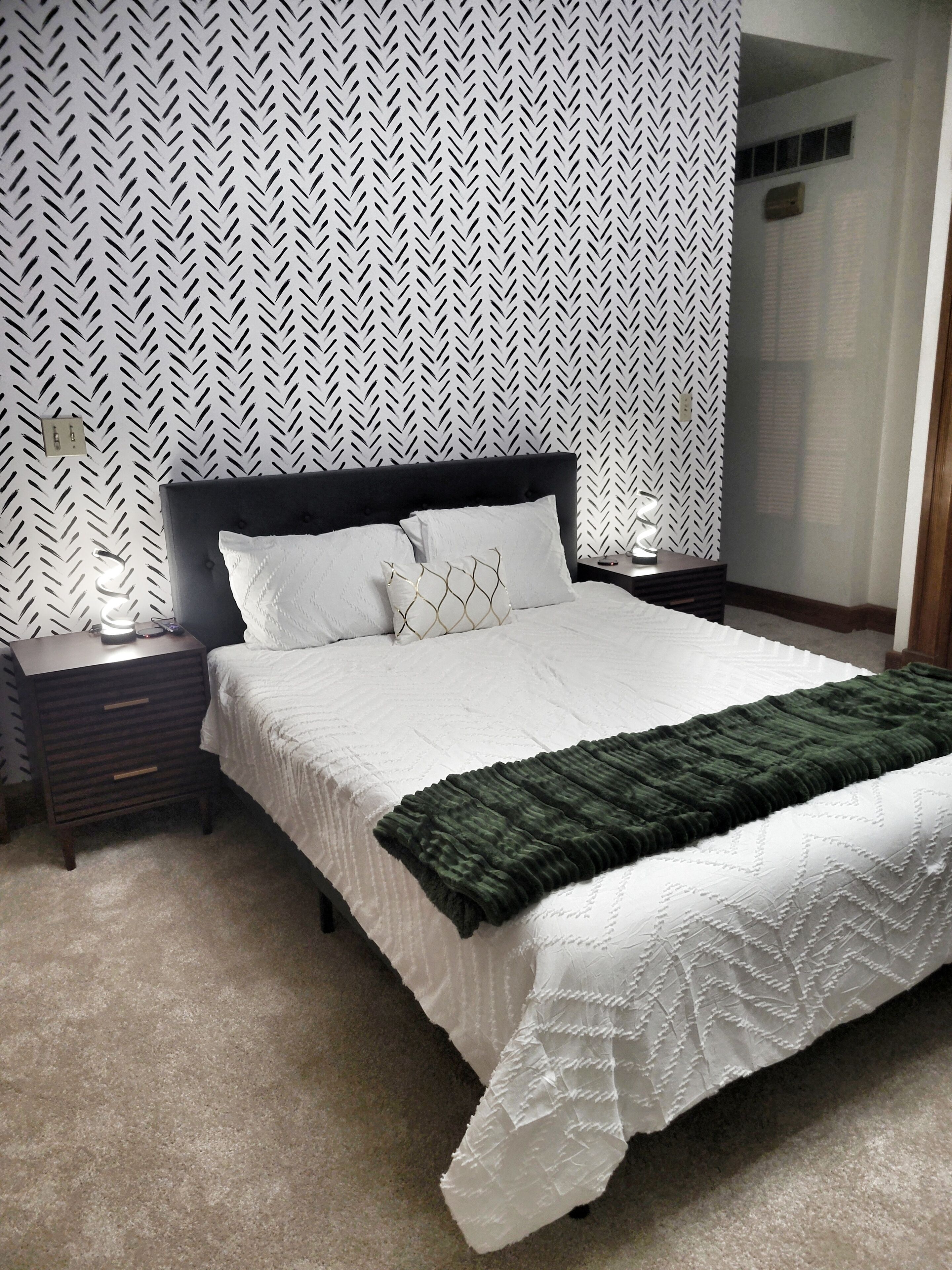 A neatly made bed with a white comforter and a green throw blanket, a dark headboard, and a nightstand with a lamp.
