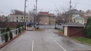 A parking lot with a white line marking the center, surrounded by a low fence and a grassy area with scattered leaves.
