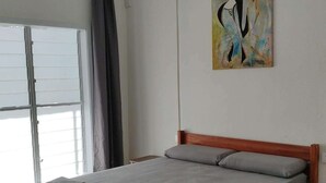 Quarto com cama de madeira, roupa de cama cinzenta, ventoinha no teto, janela com cortinas e um quadro na parede.