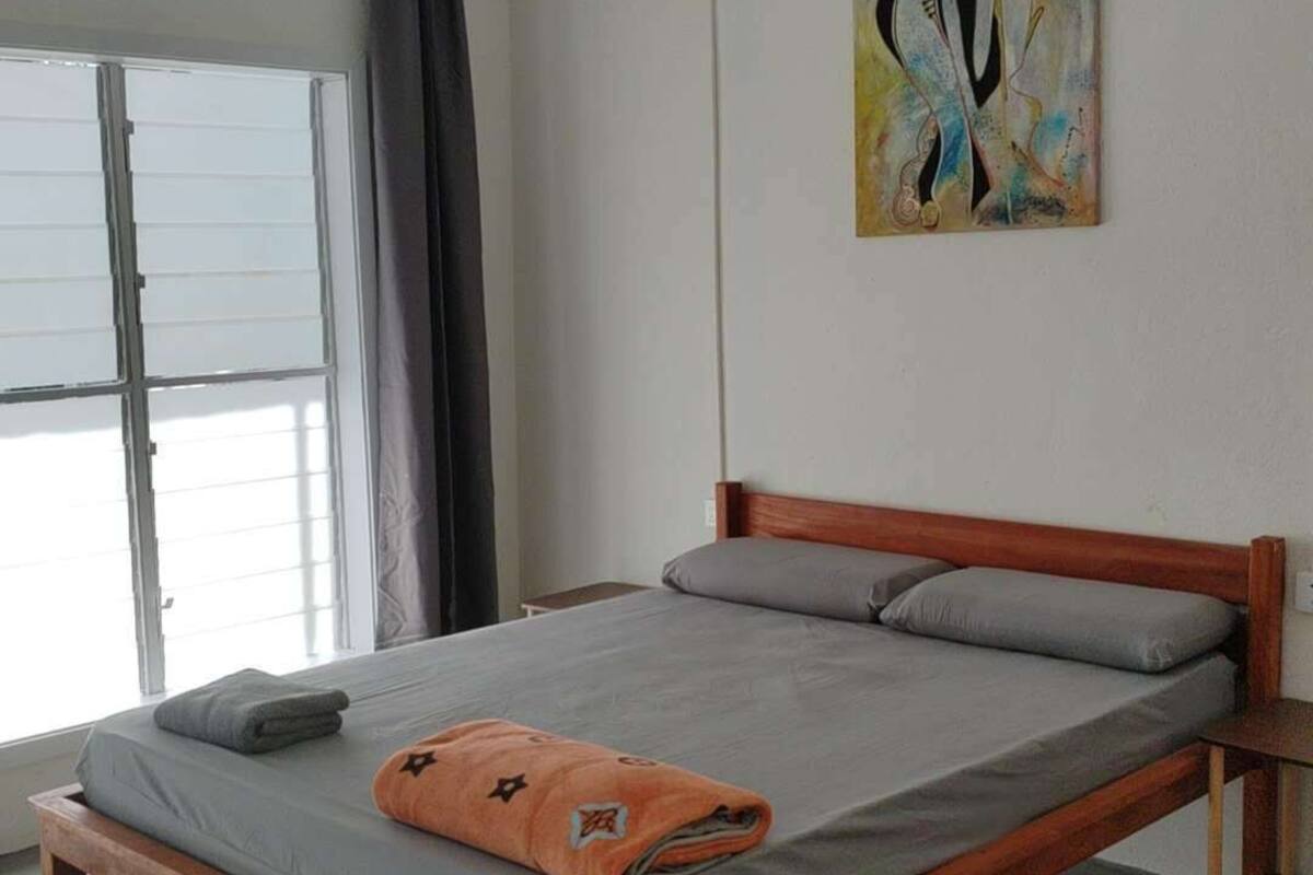 Une chambre à coucher avec un lit en bois, une literie grise, un ventilateur de plafond, une fenêtre avec des rideaux et un tableau au mur.