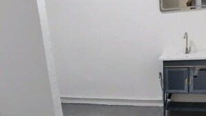 Quarto pequeno e simples, com parede branca, chão cinzento e uma pia com espelho acima.