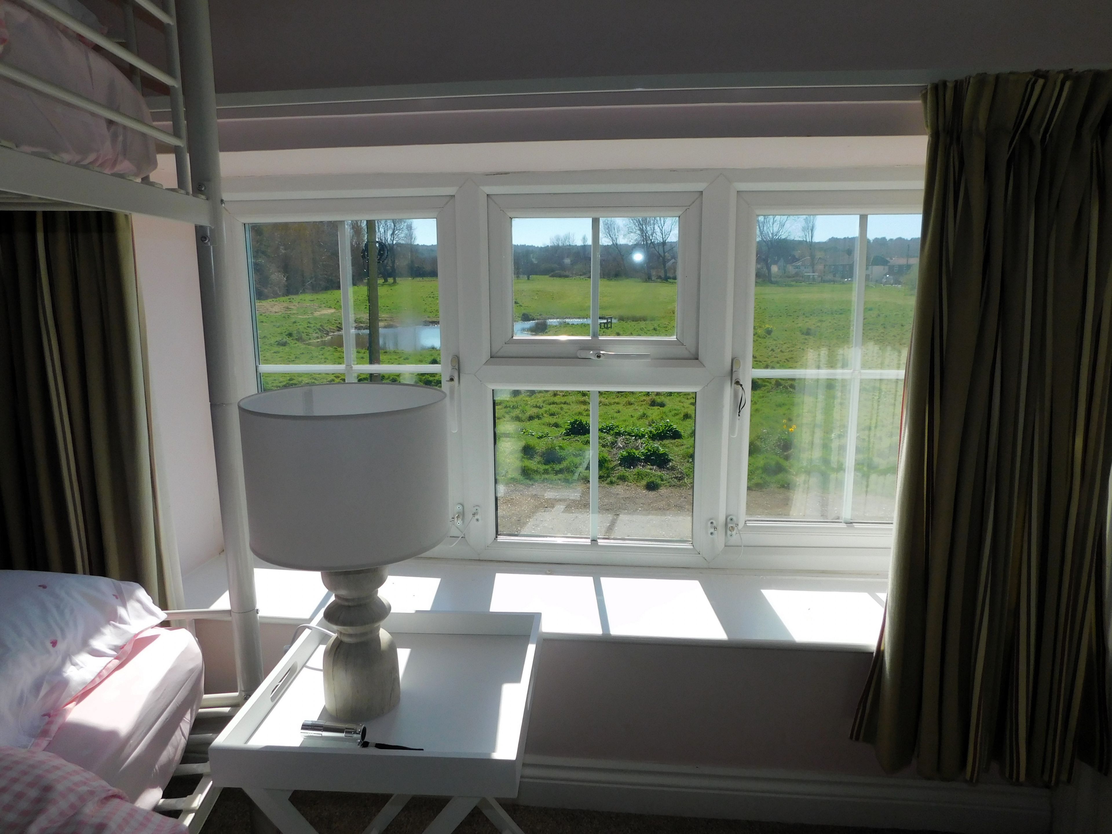 A room with a bed, a table with a lamp, and a view of a green landscape through large windows.