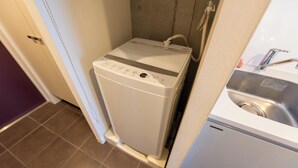 A compact, white washing machine in a small bathroom with a sink and a door leading to another room.
