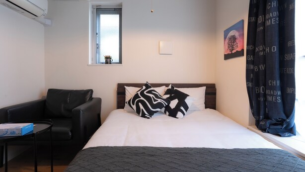 A hotel room with a bed, a black leather chair, a small table, a window with a view, and a painting on the wall.