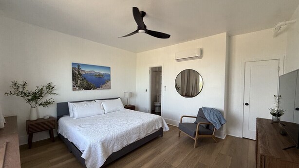 Ein Schlafzimmer mit Bett, Stuhl, Schreibtisch, Deckenventilator, Gemälde und Spiegel.