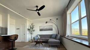 Ein modernes Wohnzimmer mit grauem Sofa, einem Couchtisch, einer Essecke mit Holztisch und -stühlen, einem Deckenventilator und einem großen Fenster mit Vorhängen.