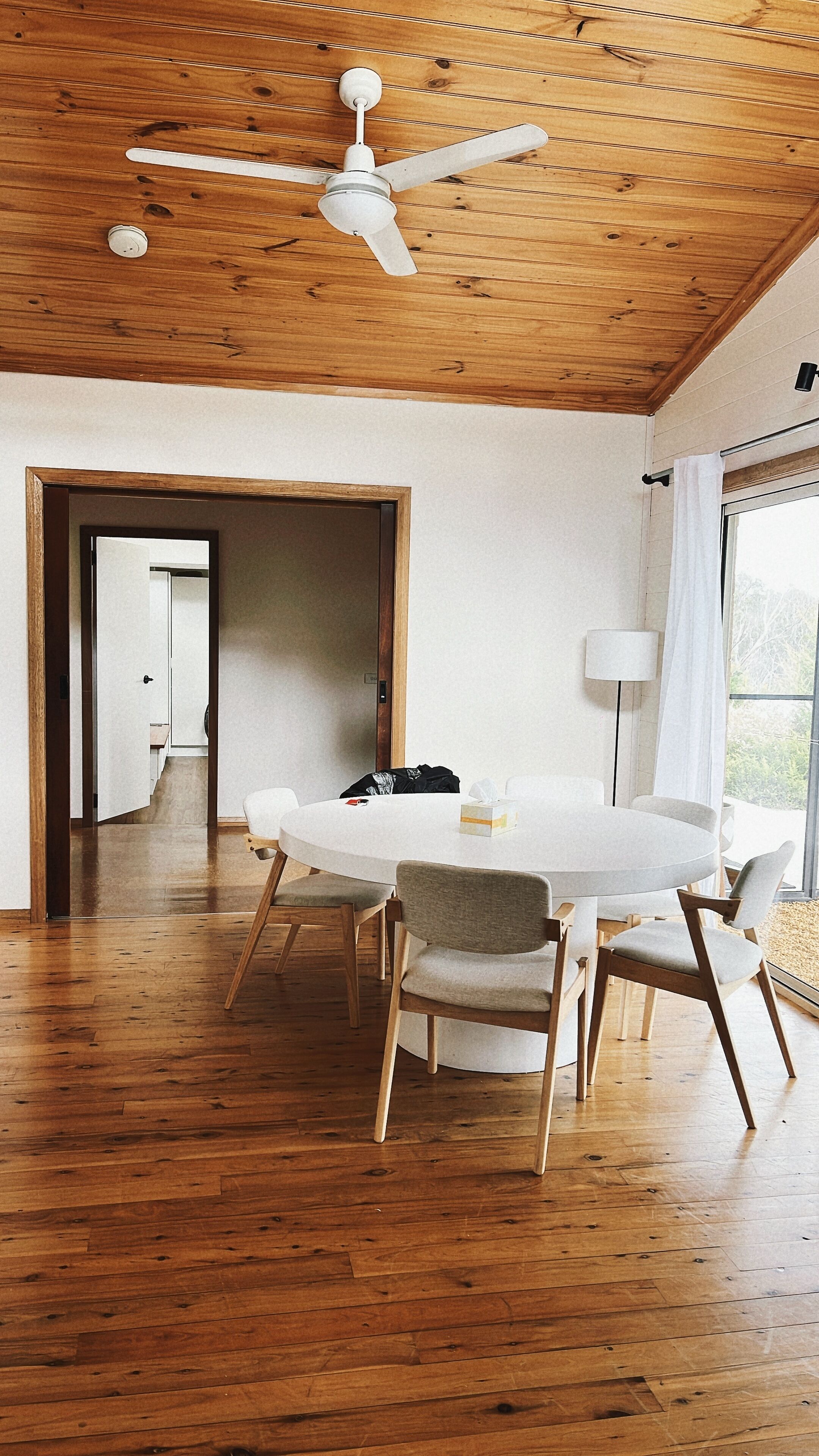 Ein geräumiges Zimmer mit Holzbalkendecke und Deckenventilator, ein runder Esstisch mit Stühlen und Blick auf einen Garten durch ein Fenster.