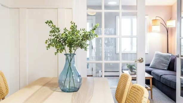 Une pièce comprenant une table à manger, des chaises en osier, un vase en verre avec des plantes vertes, un canapé et un lampadaire.