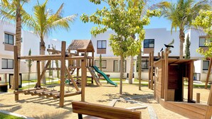 A playground with a wooden slide and swings, surrounded by palm trees and a bench. - Palmilla Sunscape  (Mazatlán)