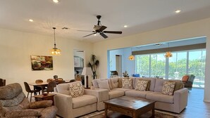 A spacious living room with a large sectional sofa, a coffee table, and a patterned rug. There is a dining area with a table and chairs, and a ceiling fan. - Golf & Gulf – Pool Home Near Beach w/ Cart (Englewood)