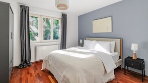 A bedroom with a bed, a nightstand, a lamp, a window with curtains, and a wall-mounted artwork.