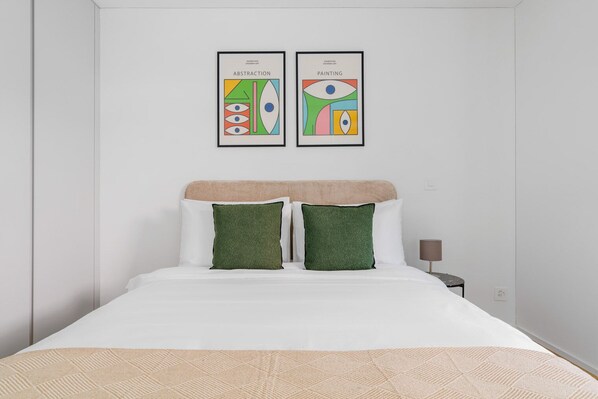 A neatly made bed with white bedding and two green pillows, a beige headboard, and two framed artworks on the wall above.
