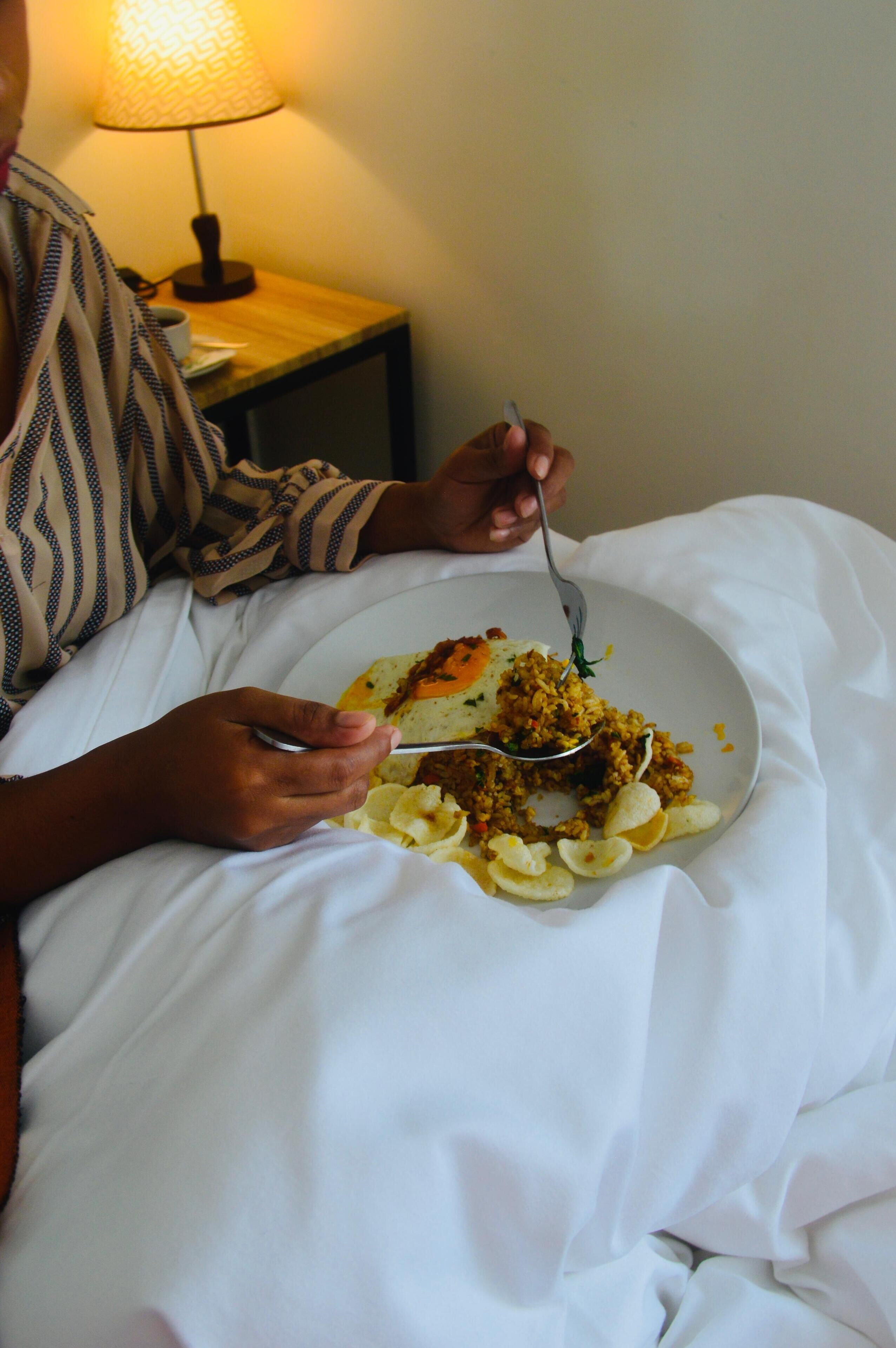 A person sitting on a bed with a plate of food, a lamp in the background, and a white pillow.