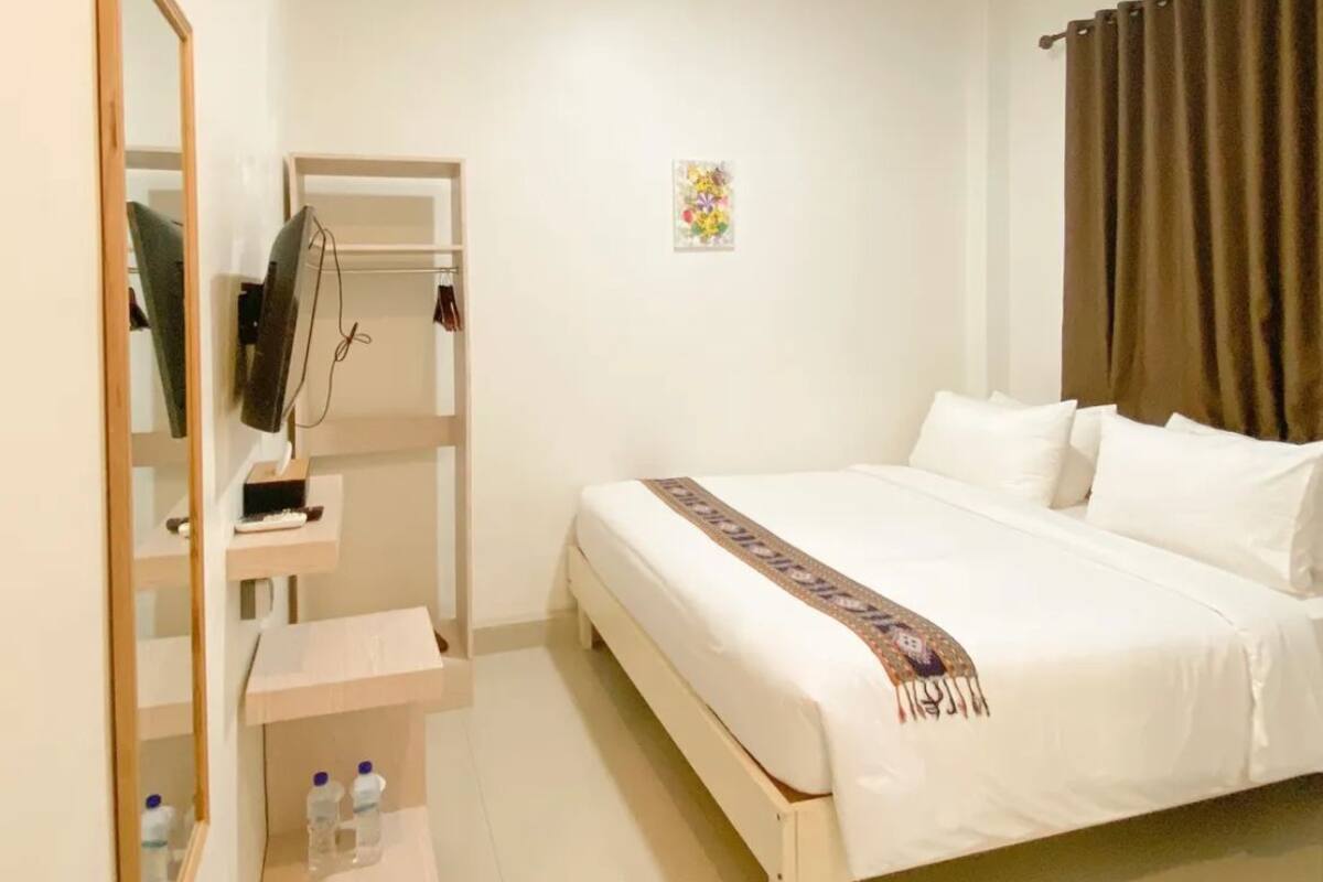 A bedroom with a bed, a TV, a mirror, a shelf with bottles, and a painting on the wall.