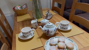 Uma mesa de jantar de madeira com uma chaleira floral, xícaras, pires e um prato de scones.