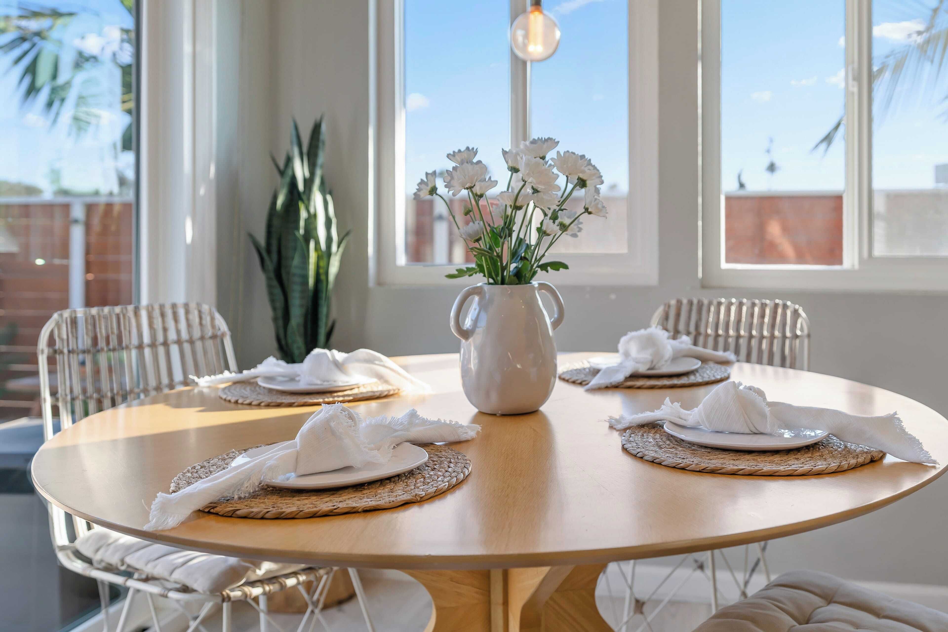 Une table de salle à manger ronde, agrémentée de sets de table blancs, de serviettes blanches et d’un pichet blanc orné de fleurs. Quatre chaises blanches sont disposées autour de la table.