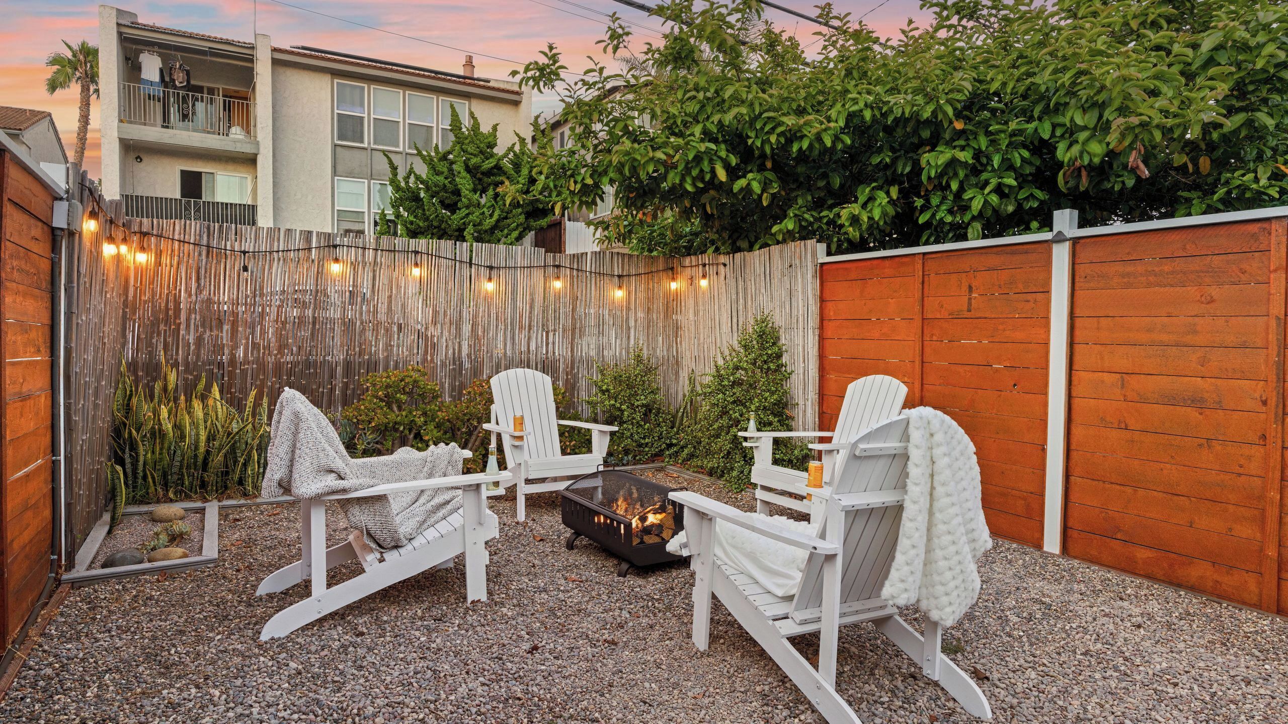 A backyard with a fire pit, white Adirondack chairs, and a wooden fence.