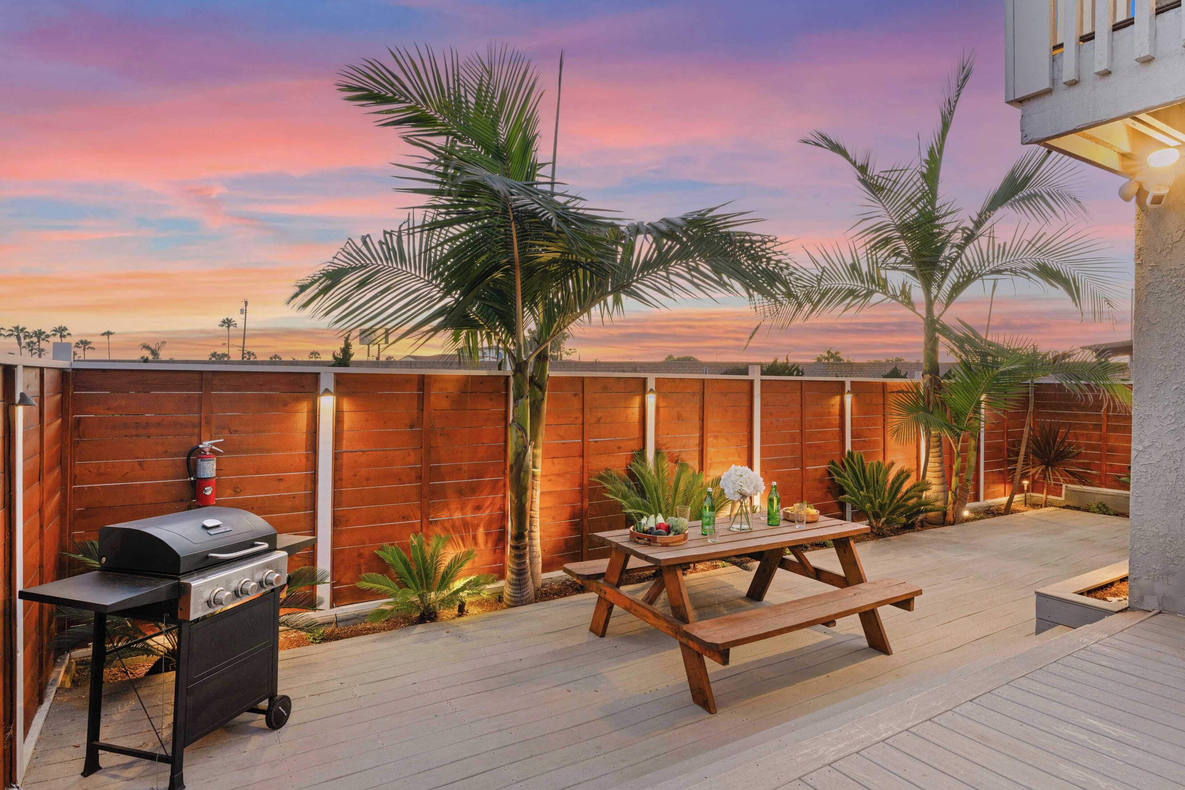 A backyard with a barbecue grill, wooden picnic table, and palm trees.