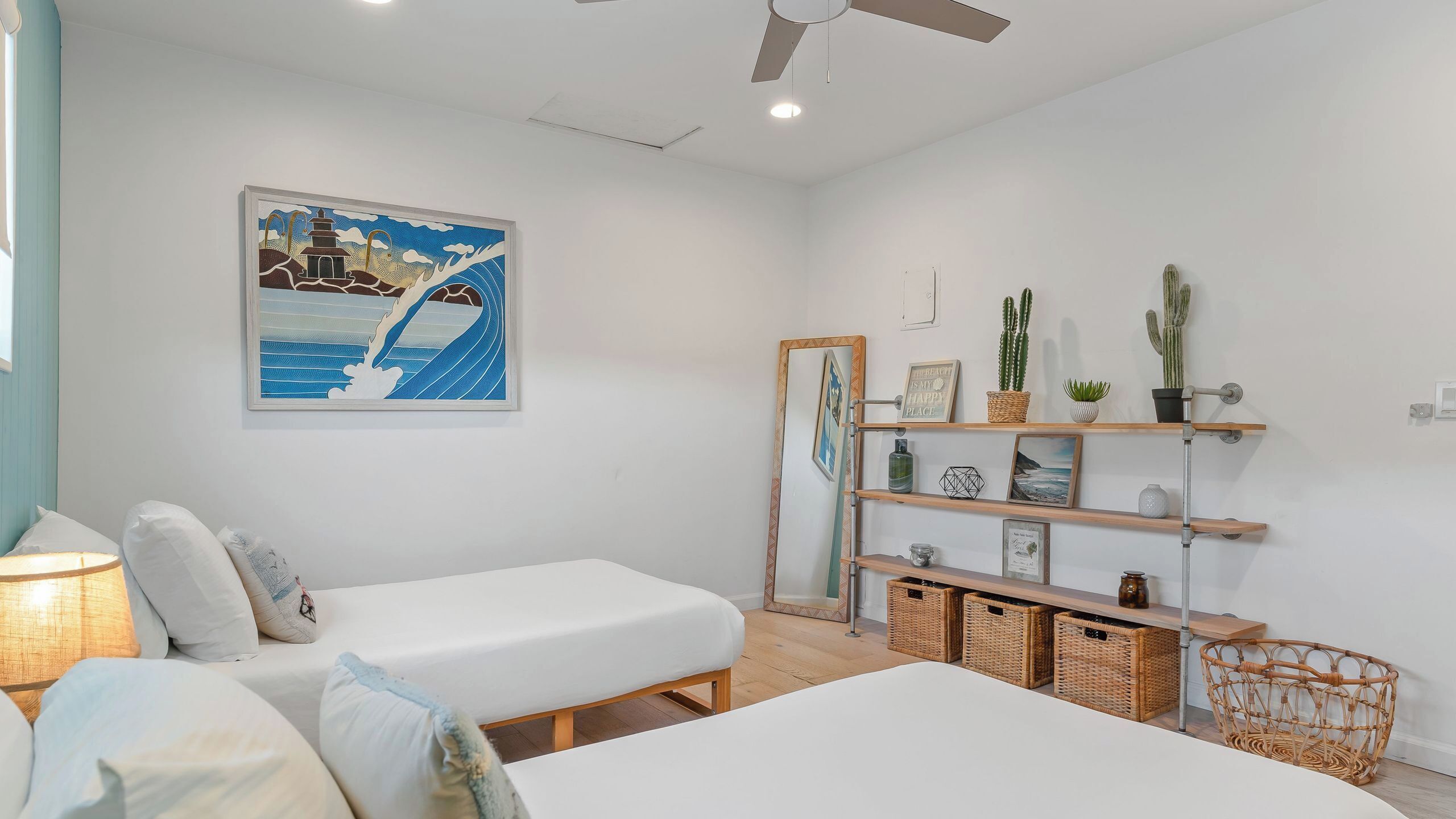 A bedroom with two beds, a ceiling fan, a painting, a shelf with plants, and a mirror.