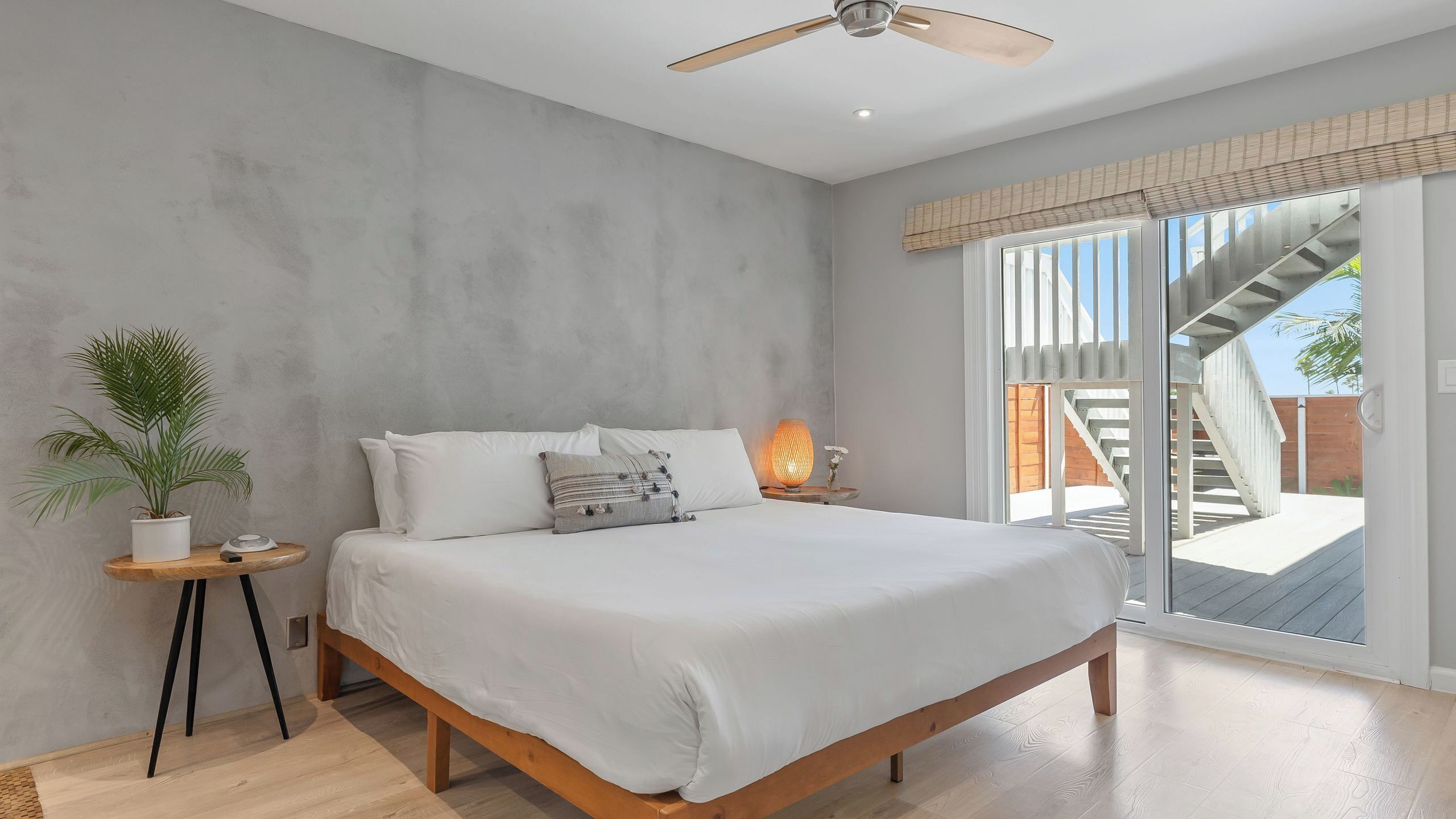A bedroom with a bed, a small bedside table, a potted plant, and a window with a view of a staircase and outdoor area.