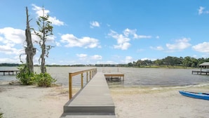 Eine Holzstege führt zu einem Steg über einem Sandstrand mit ruhigem Wasser.