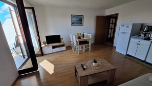 A living room with a TV, a dining area with a table and chairs, a kitchen with a fridge, and a large window. - A apartment with balcony and the sea view (Seget Vranjica)