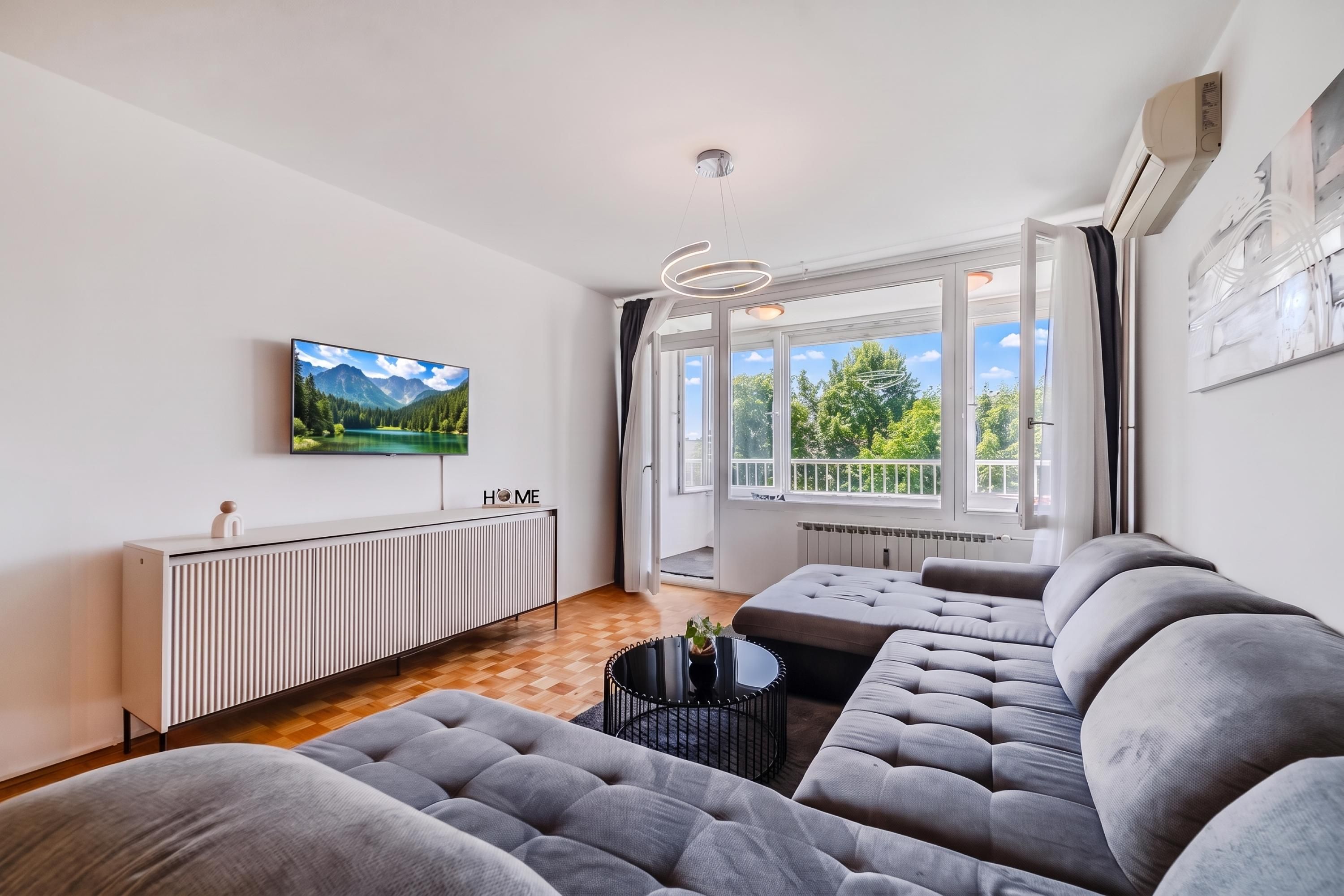 A modern living room with a large flat-screen TV, a sofa, a coffee table, and a view of greenery through large windows.