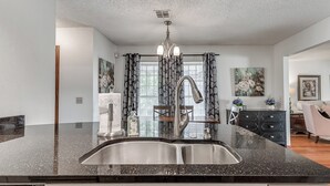 A kitchen with a black granite countertop, stainless steel sink, and a modern faucet. There is a dining area with a wooden table and chairs, and a painting on the wall.