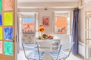 A room with a table set for two, a window with a view of buildings, and a door with colorful glass panels. - Old Castle Apartment, Menton, France (Menton)