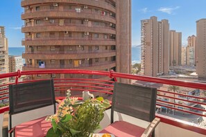 A balcony with a red railing, a table with a potted plant, and a view of a coastal cityscape with high-rise buildings.