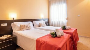 Habitación de hotel con dos camas, cada una con ropa blanca y una colcha roja. Hay dos mesitas de noche, con lámparas y una pequeña planta sobre una de las camas.