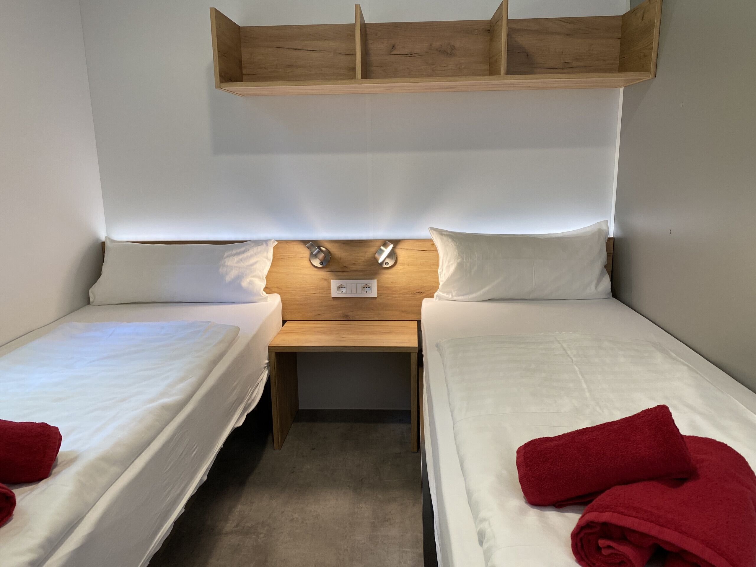 A small, single-room hotel with two single beds, a wooden headboard, bedside lamps, and a power outlet.