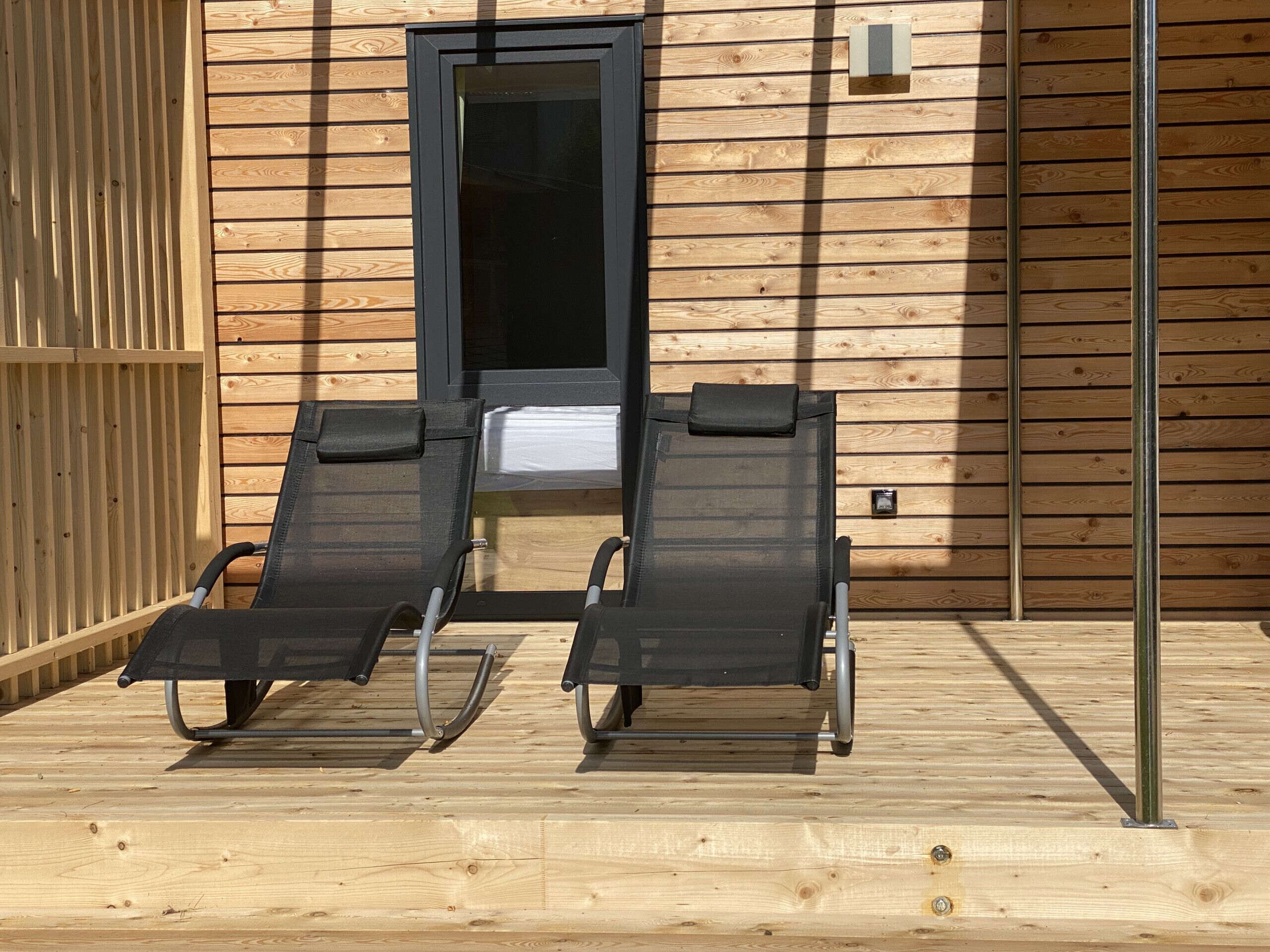Two reclining chairs with metal frames and black cushions, positioned on a wooden deck in front of a wooden building with a black-framed door and a small window.