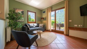 A living room with a sofa, armchair, and a small table. There is a window with a view of greenery and mountains.