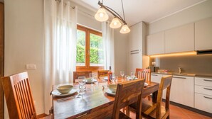 A dining area with a wooden table set for four, surrounded by chairs. A window with curtains allows natural light to enter.