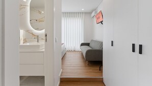 A modern hotel room with a marble bathroom vanity, a grey sofa, and a large window with white curtains. - GuestReady - Tutti Frutti Studio - Cherry (Porto)