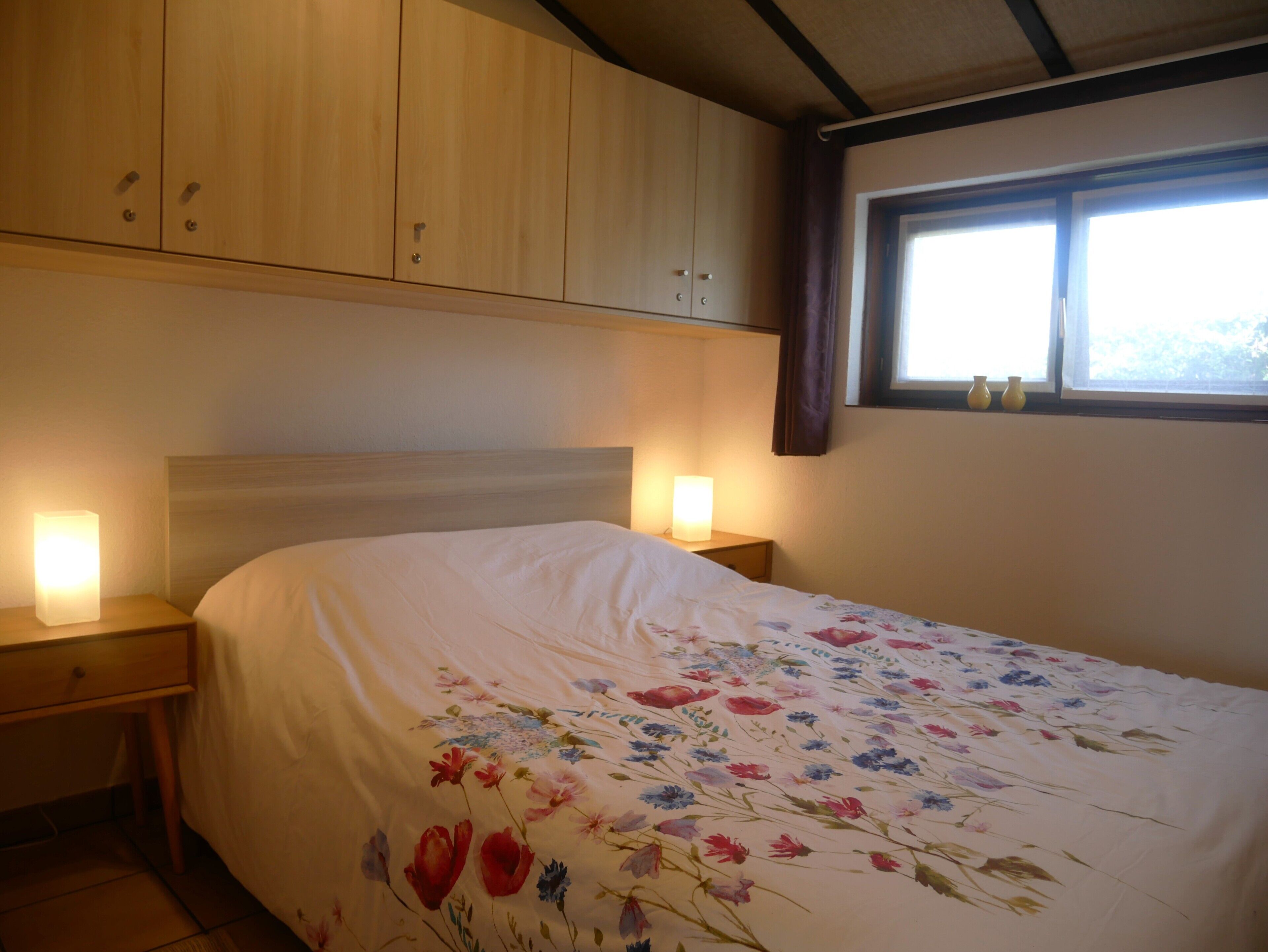 Een slaapkamer met een bed, houten kasten, een nachtkastje, een raam en een lamp.