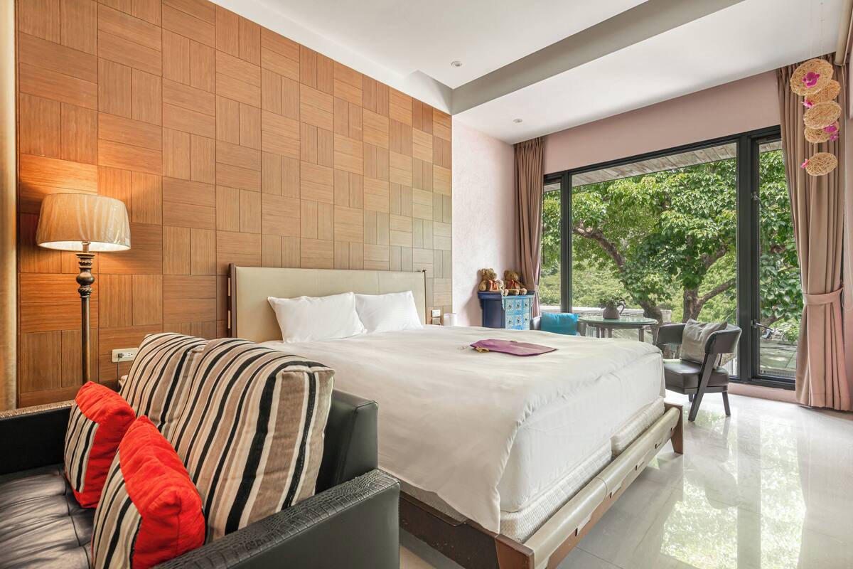 Una habitación de hotel moderna con una cama grande, un sofá con cojines a rayas, una mesa con sillas y vistas al exterior a zonas verdes.