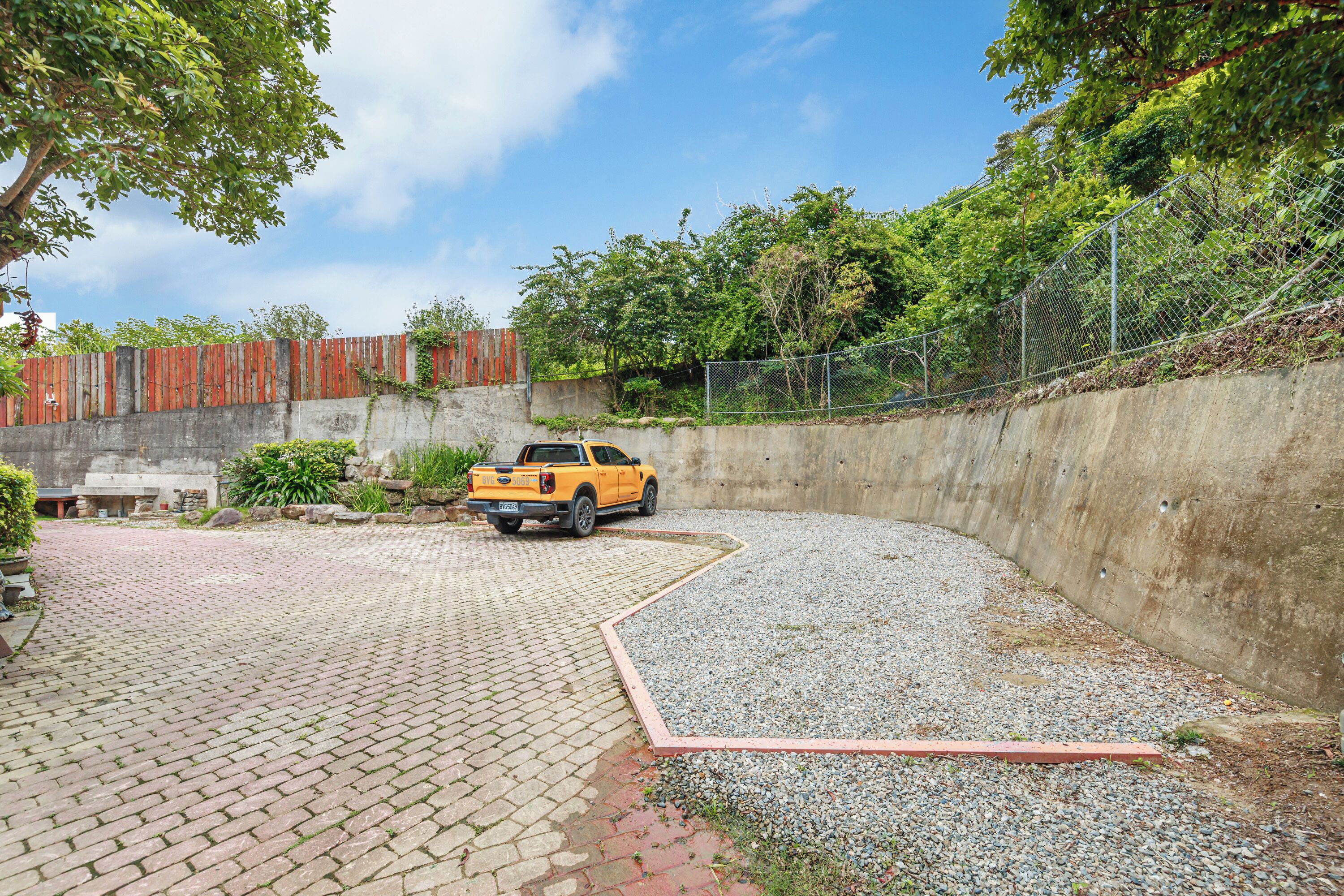 A yellow pickup truck parked in a gravel area with a paved walkway and a concrete wall.
