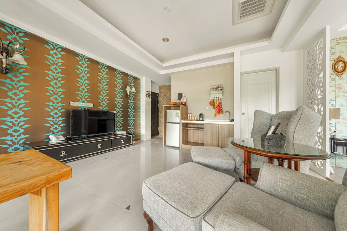 Una sala de estar moderna con televisor, sofá y reposapiés. Se observa un área de cocina al fondo, con refrigerador y pileta.