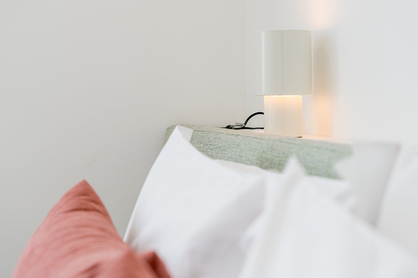 Ein ordentlich bezogenes Bett mit weißen und rosafarbenen Kissen, eine weiße Wand und eine Wandlampe.