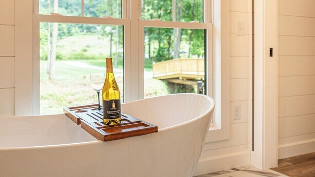 Ein modernes Badezimmer mit einer freistehenden Badewanne, einem Fenster mit Blick auf Grünflächen und einer Weinflasche am Rand der Wanne.