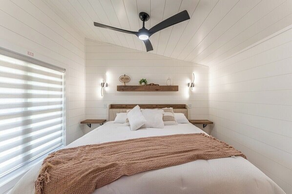 Ein Schlafzimmer mit Bett, Deckenventilator, Fenster mit Jalousien und Wandleuchten.