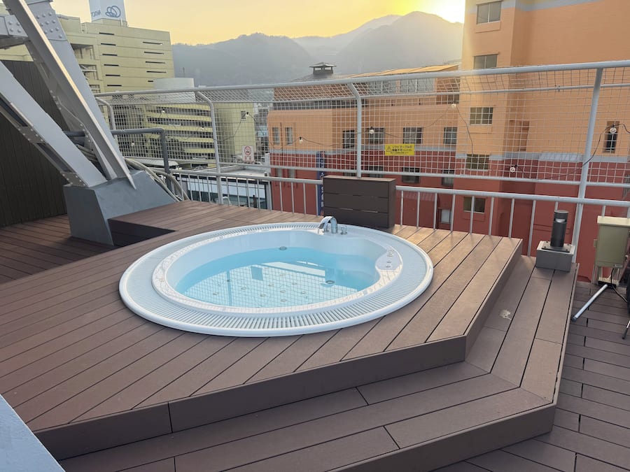 Terraço com banheira de hidromassagem, deck de madeira e vista para edifícios e montanhas.