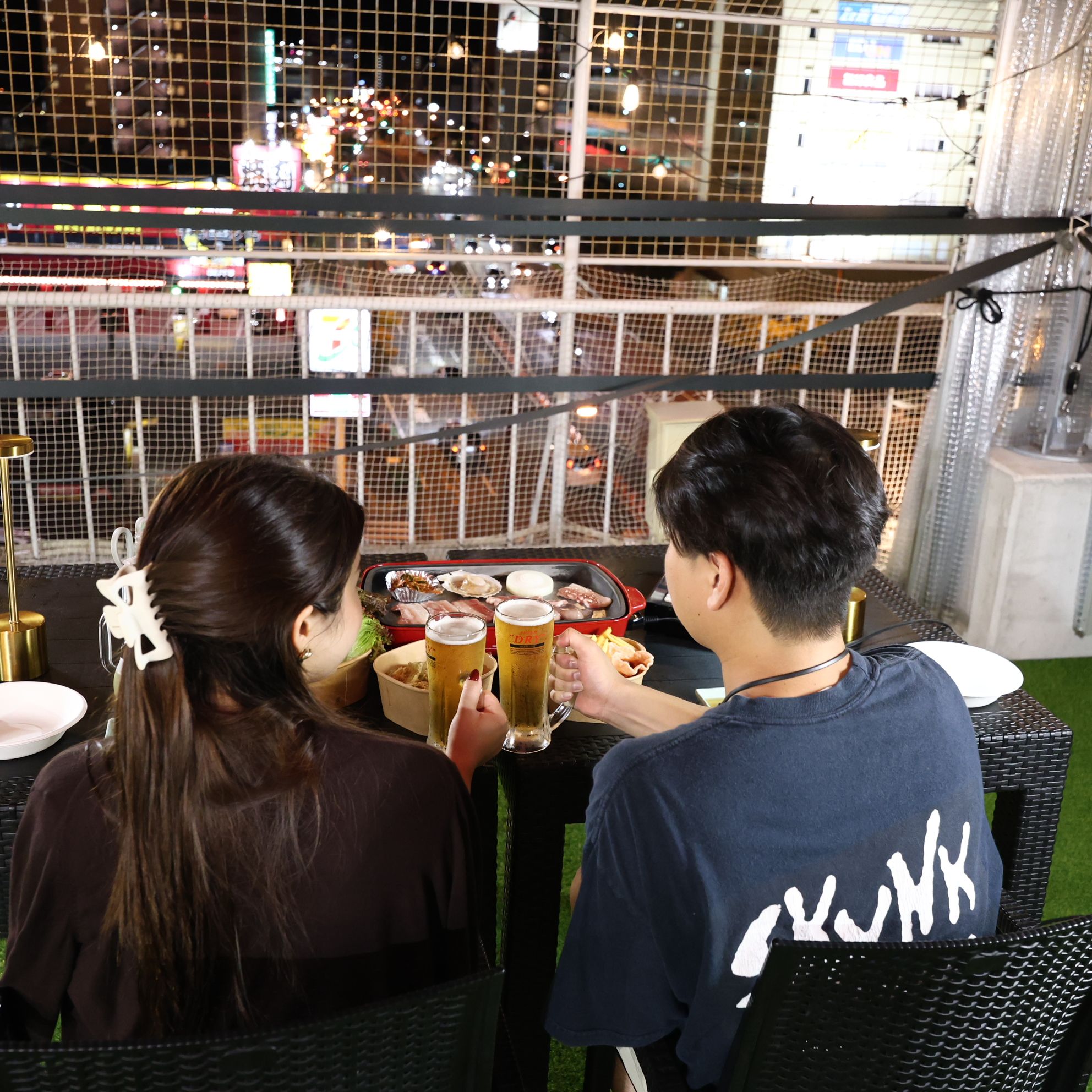 Two people seated at an outdoor table with a view of a cityscape through a wire mesh.