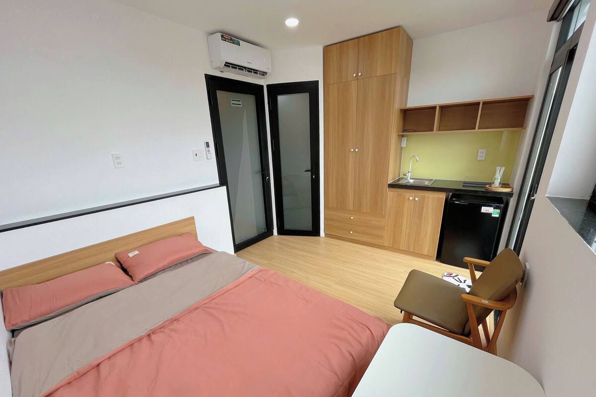 A compact room with a bed, a small desk, a chair, a kitchenette, and a wardrobe.